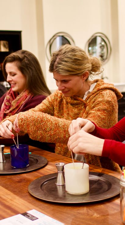 Group of women at an indoor candle-making workshop dipping wicks into glass jars around a communal wooden table in a cozy craft studio