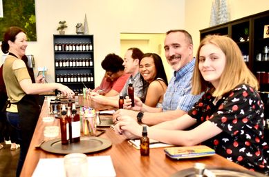 Smiling instructor leads a small group at an indoor DIY scent-making workshop, participants seated at a wooden counter mixing fragrances and holding amber bottles in a cozy craft studio.