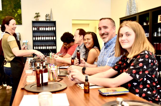 Smiling instructor leads a small group at an indoor DIY scent-making workshop, participants seated at a wooden counter mixing fragrances and holding amber bottles in a cozy craft studio.