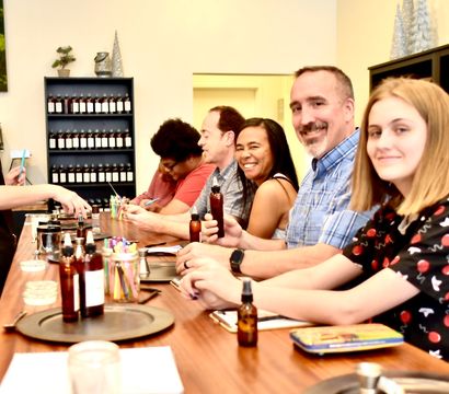 Smiling instructor leads a small group at an indoor DIY scent-making workshop, participants seated at a wooden counter mixing fragrances and holding amber bottles in a cozy craft studio.