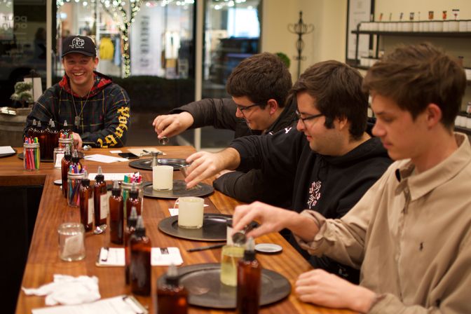 Group of four people in a cozy craft studio pouring fragrances into candle jars at a long wooden table lined with amber bottles and tools.