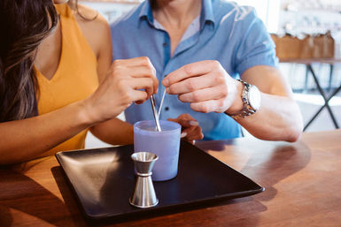 Hands-on DIY candle-making: two people stirring melted wax in a purple jar, with a metal measuring jigger and black tray on a wooden table.