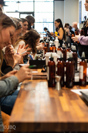 Hands-on fragrance workshop at a long wooden table, people sampling scents from amber spray bottles in a bright studio
