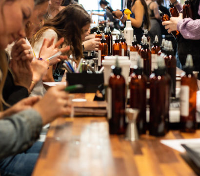 Hands-on fragrance workshop at a long wooden table, people sampling scents from amber spray bottles in a bright studio
