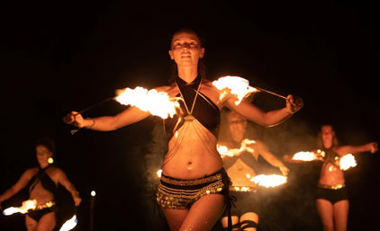 Fiery night performance: central dancer twirls flaming torches in a sequined costume with other fire dancers silhouetted against a dark background.