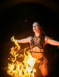 Nighttime fire performer in strappy black costume and gold chains kneeling with arms outstretched as bright orange flames swirl in front against a dark background