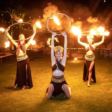 Three fire dancers in black-and-gold costumes performing with flaming hoops at an outdoor nighttime luau-style show on grass, surrounded by tiki torches and string lights.