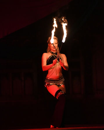 Female fire performer on a dim red-lit stage holding crossed flaming torches above her while wearing a sparkling belly-dance style costume.