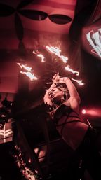 Fire performer spinning a multi-pronged flaming fan overhead on an indoor nightclub stage, striking a dramatic pose under moody red lighting.