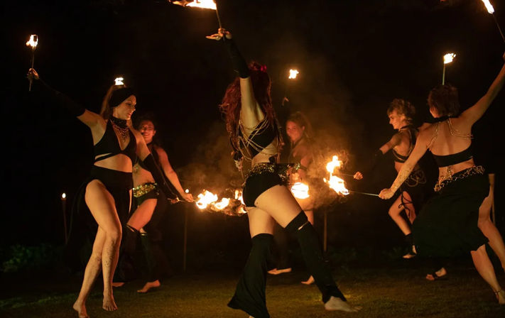 Energetic nighttime outdoor fire performance with costumed dancers twirling flaming torches and fire props over a grassy lawn
