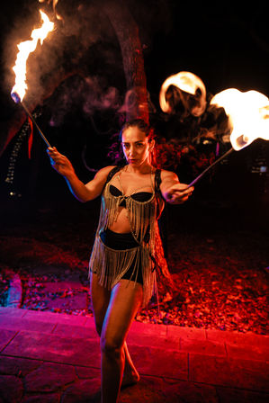 Female fire dancer twirling flaming torches in an outdoor nighttime performance, wearing a fringe chain costume under red and purple dramatic lighting.