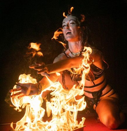 Fire dancer kneeling in a nighttime stage performance, swirling bright orange flames around their arms and hands in a dramatic, mesmerizing fire dance.