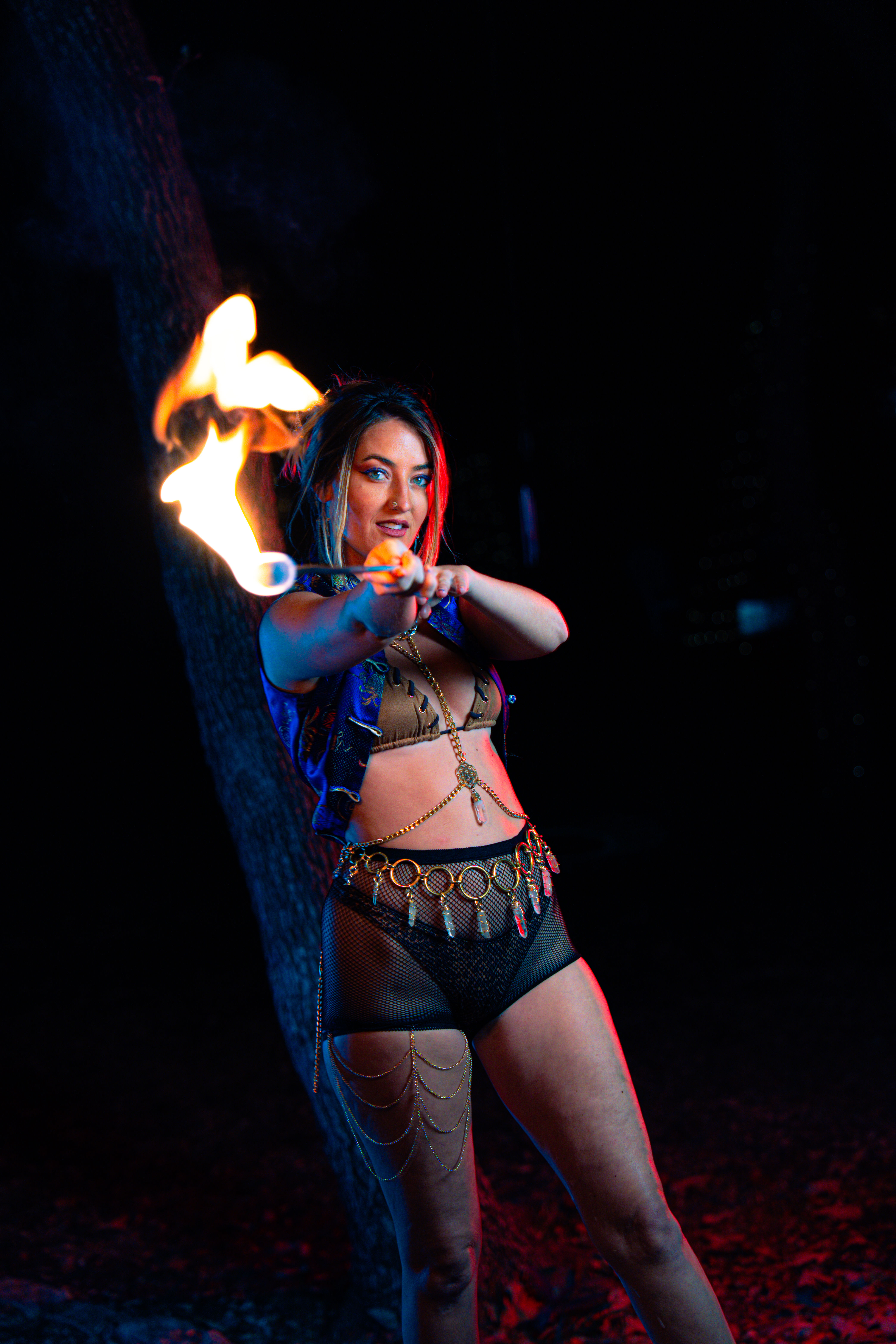 Fire performer outdoors at night holding a flaming torch toward the camera, wearing a festival-style costume with gold coin belt, mesh shorts and body chains, lit by dramatic blue and red lighting against a dark, tree-lined background.