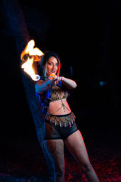 Fire performer outdoors at night holding a flaming torch toward the camera, wearing a festival-style costume with gold coin belt, mesh shorts and body chains, lit by dramatic blue and red lighting against a dark, tree-lined background.