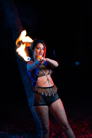 Fire performer outdoors at night holding a flaming torch toward the camera, wearing a festival-style costume with gold coin belt, mesh shorts and body chains, lit by dramatic blue and red lighting against a dark, tree-lined background.
