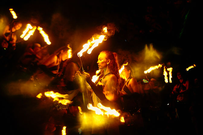 Female fire dancer twirling flaming torches in a group performance at an outdoor nighttime festival, glowing orange flames and dramatic motion blur