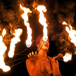 Fire performer twirling multiple flaming wicks around face and hands in a dramatic night outdoor fire-dancing show, bright orange flames and smoke against a dark background.