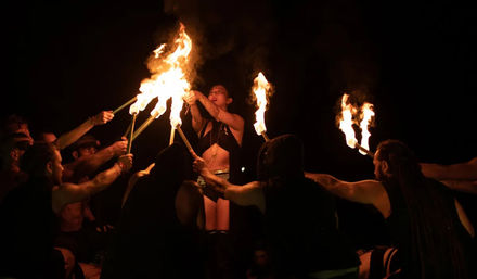 Nighttime fire performance: a central performer in stage costume raises flaming torches while a circle of people hold out burning sticks, dramatic orange flames lighting faces against a dark background.