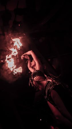 Fire performer in a sequined costume twirls flaming torches overhead on a smoky, red-lit stage — dramatic fire dance.