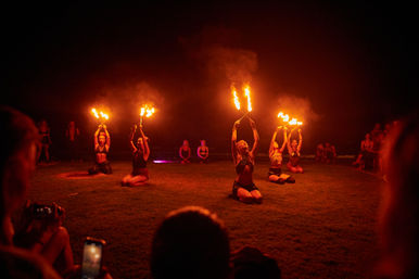 Fire dancers performing outdoors at night on a grassy lawn — five kneeling performers holding flaming torches while a silhouetted audience watches.