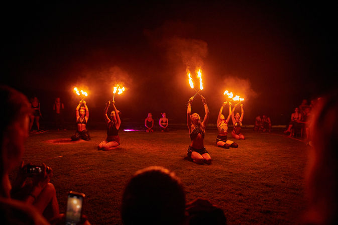 Fire dancers performing outdoors at night on a grassy lawn — five kneeling performers holding flaming torches while a silhouetted audience watches.