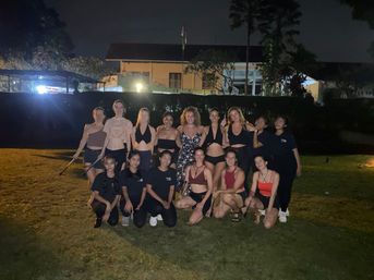 Around 16 women in activewear and swimsuits smiling for a nighttime group photo on a lit grassy lawn in front of a building and trees.