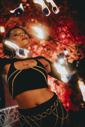 Fire performer in a black crop top with gold chain harness manipulating multiple flaming wands against a warm red floral backdrop at a nighttime event.
