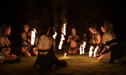 Six fire performers kneel in a circle on grass at night, each holding flaming torches in a synchronized outdoor fire dance with warm glowing flames.