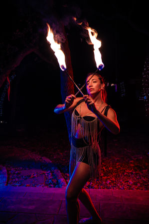 Fire dancer performing outdoors at night, crossing two flaming torches under dramatic red and blue lighting while wearing a fringed sequined costume