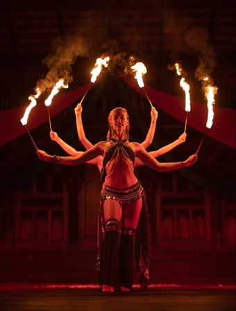 Fire performer on a dark stage holding six flaming torches in red theatrical lighting, creating a multi‑armed illusion with smoke and tribal-style costume.