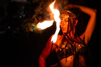 Fire performer in beaded costume twirling a flaming torch at a nighttime outdoor show, orange flames and smoke illuminating her face