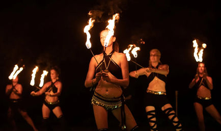Nighttime fire performance with five dancers in black-and-gold costumes spinning flaming torches, center performer holding two blazing wicks