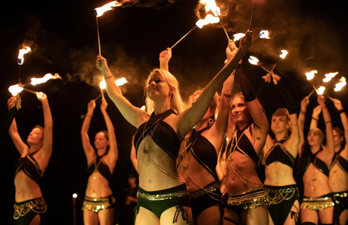 Group of synchronized fire dancers in black and gold costumes raising flaming torches overhead during a nighttime outdoor performance, glowing flames and drifting smoke against a dark background.