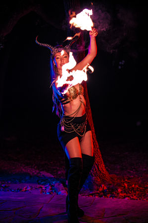 Nighttime outdoor fire performer wearing a horned headpiece and gold-sequined bra, twirling bright flames amid purple-red lighting, chain-adorned skirt and thigh-high boots by a tree.