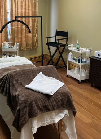 Cozy spa treatment room with a massage table draped in a brown blanket and folded white towels, a white rolling cart stocked with skincare bottles and supplies, a black director-style chair, an overhead magnifying lamp, hardwood floor and gold curtains.