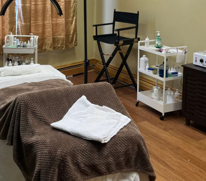 Cozy spa treatment room with a massage table draped in a brown blanket and folded white towels, a white rolling cart stocked with skincare bottles and supplies, a black director-style chair, an overhead magnifying lamp, hardwood floor and gold curtains.