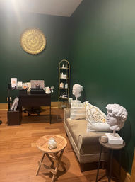 Cozy waiting area with deep green walls, gold circular wall art, velvet beige loveseat with patterned pillows, two white classical bust sculptures on side tables, small wooden coffee table with jar, dark wood desk and shelving on hardwood floor.