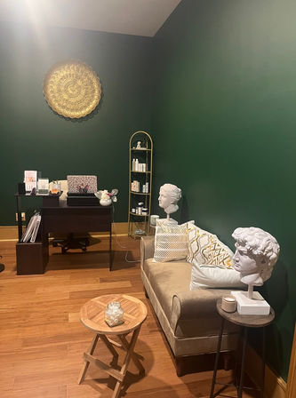 Cozy waiting area with deep green walls, gold circular wall art, velvet beige loveseat with patterned pillows, two white classical bust sculptures on side tables, small wooden coffee table with jar, dark wood desk and shelving on hardwood floor.