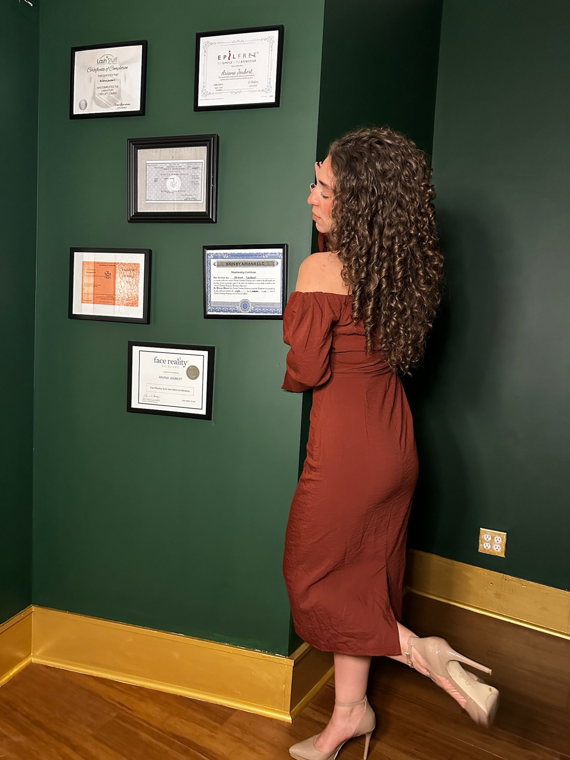 Woman with long curly hair in a rust off-shoulder midi dress and nude heels standing on tiptoe, admiring framed certificates on a dark green wall with gold baseboard in a boutique studio interior.