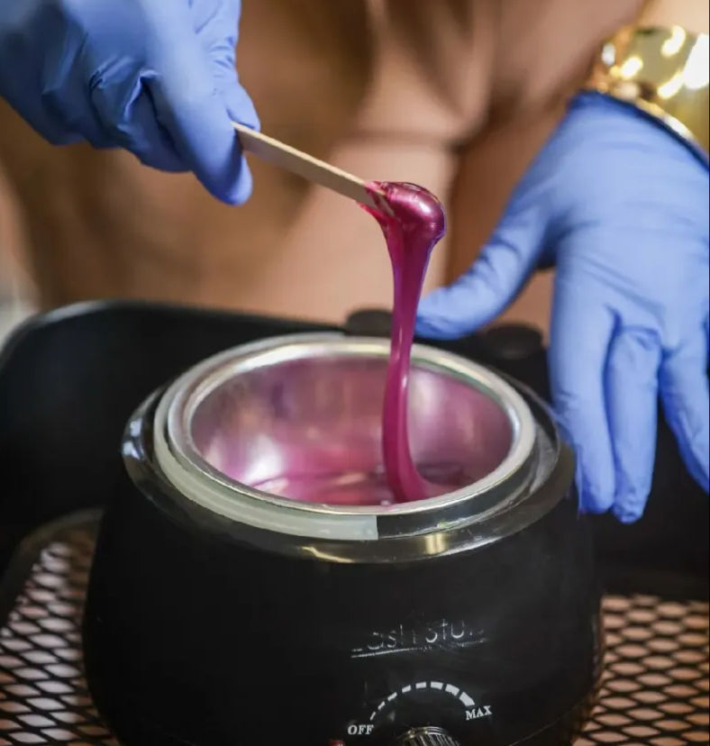 Close-up of gloved hands lifting gooey pink depilatory wax from a black wax warmer with a wooden spatula in a salon setting.