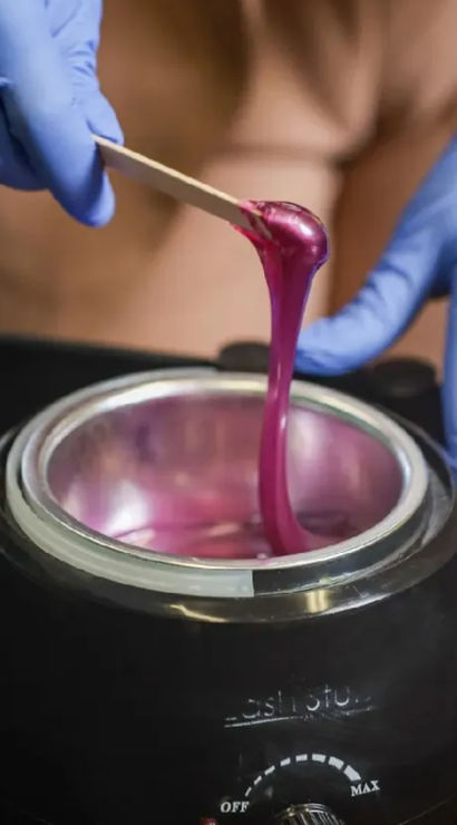 Close-up of gloved hands lifting gooey pink depilatory wax from a black wax warmer with a wooden spatula in a salon setting.