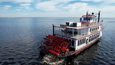 Red-and-white paddlewheel riverboat cruising on a wide calm river under a bright blue sky with scattered clouds