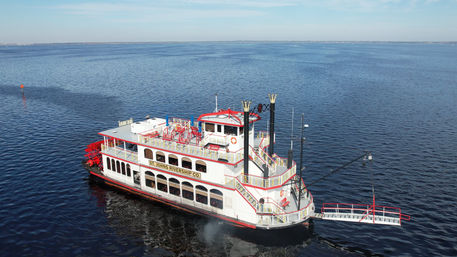 White-and-red paddlewheel riverboat sightseeing cruise on calm blue water with multi-level open decks, twin smokestacks, and an extended boarding ramp