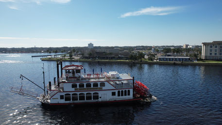 White-and-red paddlewheel riverboat gliding on a sunlit river past a palm-lined waterfront and low-rise city buildings.
