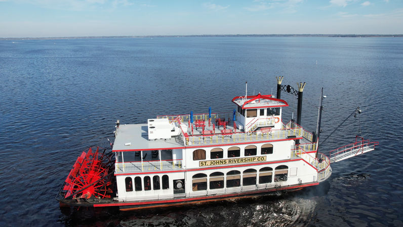 Aerial view of a white-and-red paddlewheel riverboat with twin smokestacks and an open upper deck cruising on a wide calm river under a clear blue sky.