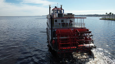Rear view of a red-and-white paddlewheel riverboat chugging along a sunny Florida river, large red paddlewheel splashing water and a palm-lined shoreline under a blue sky.