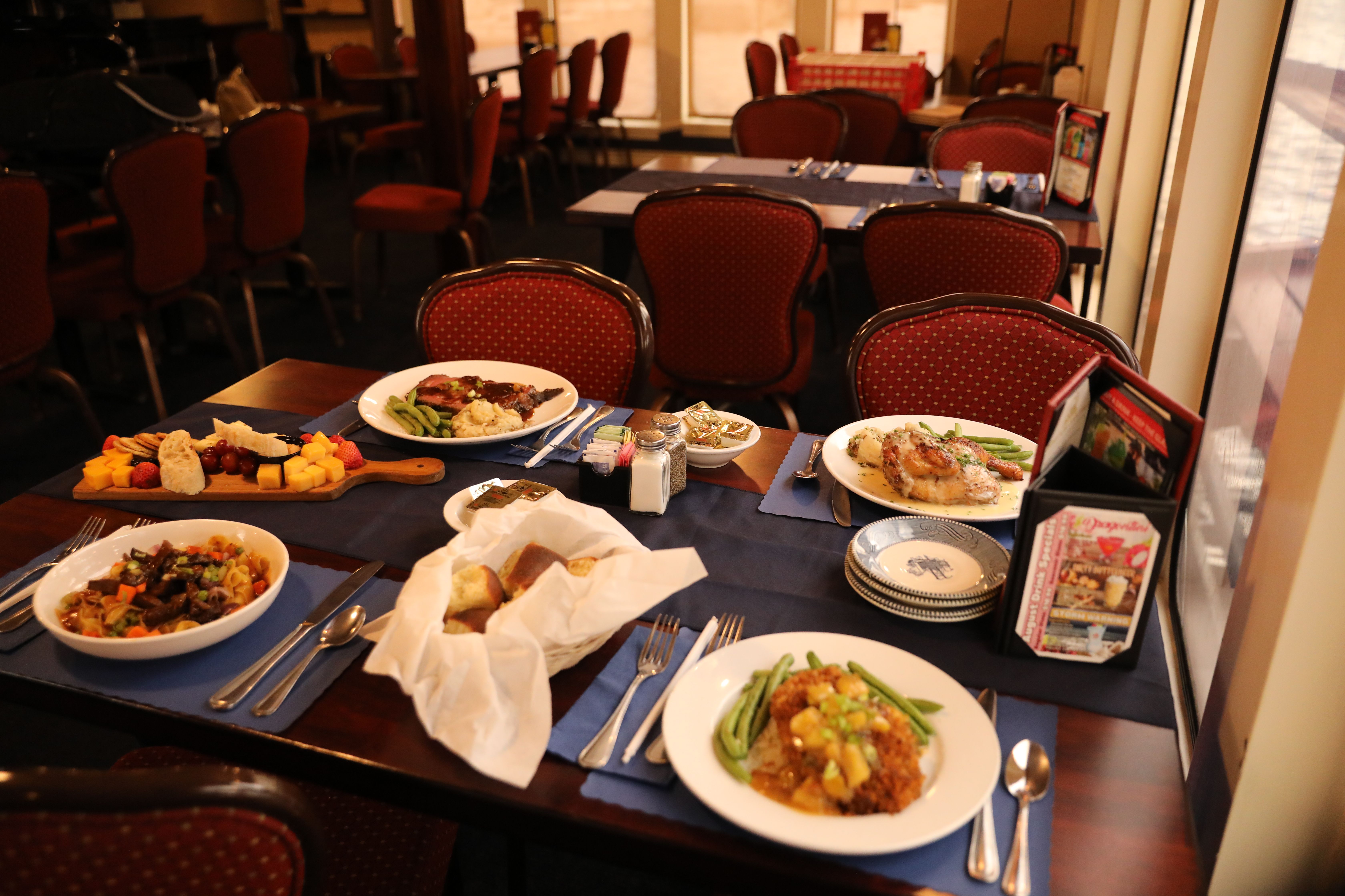 Cozy bistro-style restaurant table set for four with an inviting spread: roasted chicken with green beans, steak with mashed potatoes, a saucy rice dish, bread basket and a cheese-and-fruit board on navy placemats.