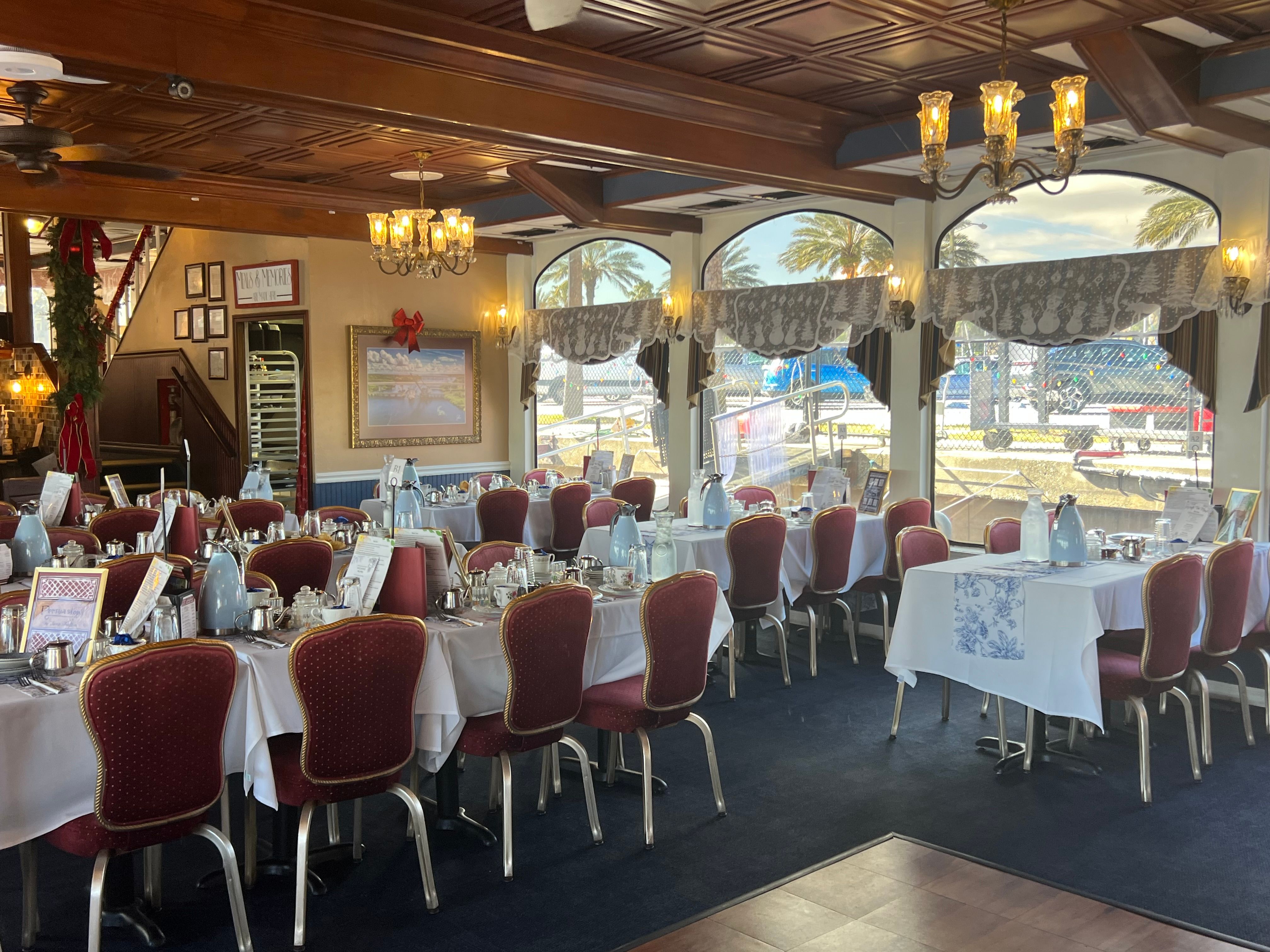 Coastal waterfront dining room set for service with white tablecloths, red upholstered chairs, chandeliers and arched windows framing palm trees and boats at the harbor.