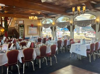 Coastal waterfront dining room set for service with white tablecloths, red upholstered chairs, chandeliers and arched windows framing palm trees and boats at the harbor.