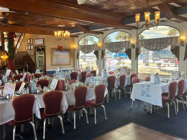 Coastal waterfront dining room set for service with white tablecloths, red upholstered chairs, chandeliers and arched windows framing palm trees and boats at the harbor.